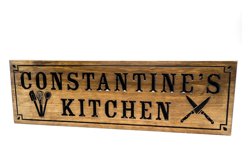 以下が含まれることがあります： 黒い文字で「CONSTANTINE'S KITCHEN」と書かれた木製の看板。看板は濃い茶色に着色され、装飾的な縁取りが施されています。木にはキッチン用品と交差したナイフが彫刻されています。