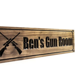 May include: A wooden sign with a dark brown stain and black lettering. The sign reads "Ben's Gun Room" and features a pair of crossed rifles.