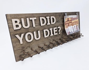 May include: A rectangular dark wood display board featuring the light-colored wooden text BUT DID YOU DIE? with a row of pegs along the bottom for hanging items and two metal clips holding a race bib with the number 1017 attached.