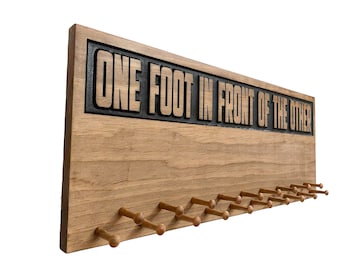 Può includere: Un porta medaglie in legno con il testo "ONE FOOT IN FRONT OF THE OTHER" inciso nella parte superiore. Il supporto ha 18 pioli in legno per appendere le medaglie.