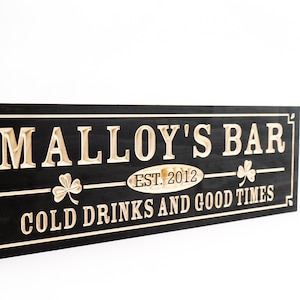 May include: Black wooden sign with white lettering that reads "Malley's Bar Est. 2012 Cold Drinks and Good Times". The sign has two shamrock designs on either side of the text.