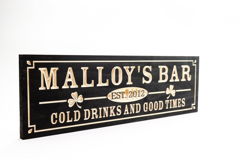May include: Black wooden sign with white lettering that reads "Malley's Bar Est. 2012 Cold Drinks and Good Times". The sign has two shamrock designs on either side of the text.