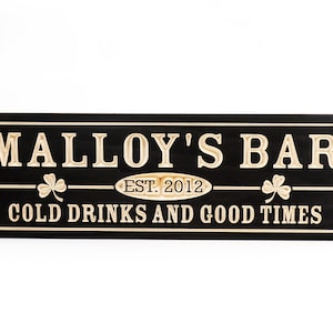 May include: Black wooden sign with gold lettering that reads "Malley's Bar Est. 2012 Cold Drinks and Good Times". The sign has two shamrock designs on either side of the text.