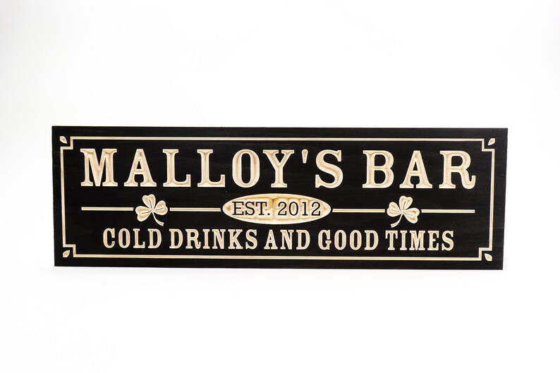 May include: Black wooden sign with gold lettering that reads "Malley's Bar Est. 2012 Cold Drinks and Good Times". The sign has two shamrock designs on either side of the text.