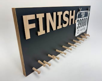 Può includere: Un cartello in legno con la scritta "FINISH" in lettere marroni chiare. Il cartello ha uno sfondo nero e diversi pioli di legno per appendere medaglie. È attaccato un pettorale con il numero 39880.