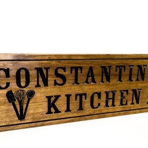 以下が含まれることがあります： 黒い文字で「CONSTANTINE'S KITCHEN」と書かれた木製の看板。看板は、黒いシルエットのキッチン用品と交差したナイフで飾られています。