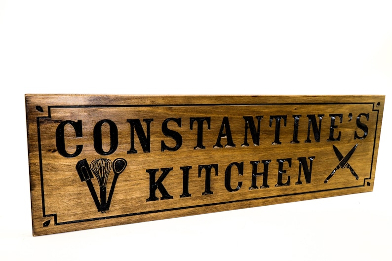 以下が含まれることがあります： 黒い文字で「CONSTANTINE'S KITCHEN」と書かれた木製の看板。看板は、黒いシルエットのキッチン用品と交差したナイフで飾られています。