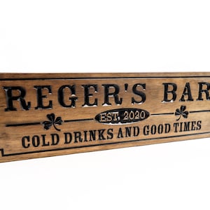 May include: A brown wooden sign with the text "Reger's Bar Est. 2020 Cold Drinks and Good Times" in black lettering. The sign has a decorative border and shamrock designs.