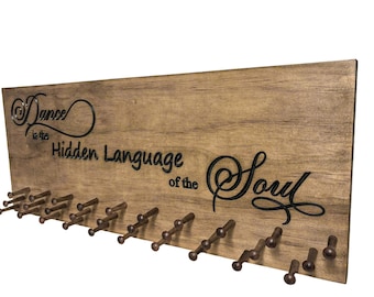 Può includere: Decorazione murale in legno con la citazione "Dance is the Hidden Language of the Soul" in caratteri neri. La tavola di legno marrone ha diversi piccoli pioli decorativi per appendere oggetti.
