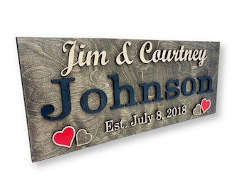 Può includere: Cartello in legno con i nomi "Jim & Courtney" e il cognome "Johnson" in un design a strati. Il cartello include la data "Est. July 8, 2018" e due forme di cuore, una rossa e una grigia.