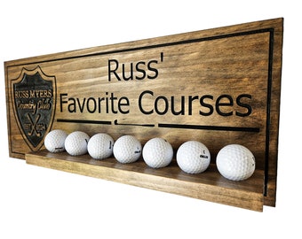 Può includere: Un cartello in legno con il testo "Russ' Favorite Courses" e un logo di club di golf. Il cartello ha uno scaffale sottostante che contiene otto palline da golf.