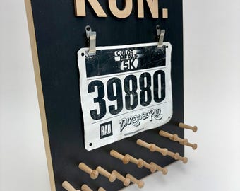 Può includere: Una bacheca nera in legno per l'esposizione dei pettorali con la scritta "RUN" in lettere di legno chiaro. La bacheca contiene un pettorale con il numero 39880 e ha pioli in legno per le medaglie.