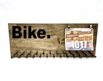 May include: A rectangular wooden wall display featuring the word Bike. in bold black lettering on the left side. The right side includes two metal clips holding a Runtoberfest race bib with the number 1017. A row of small wooden pegs lines the bottom.