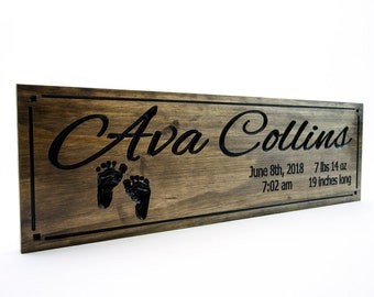 Può includere: Un cartello in legno marrone scuro con un bordo nero inciso. Il cartello recita "Ava Collins" in un carattere corsivo con due piccole impronte di piedi sotto il nome. La data "June 8th, 2018" e l'ora "7:02 am" sono incise sotto le impronte di piedi, insieme al peso e alla lunghezza del bambino: "7 lbs 14 oz, 19 inches long".
