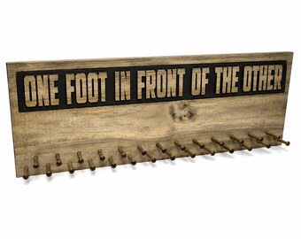 May include: A rectangular wooden wall-mounted display rack featuring a natural light brown wood grain finish. The top section displays the text ONE FOOT IN FRONT OF THE OTHER in raised wooden letters against a black background. It has sixteen hooks.