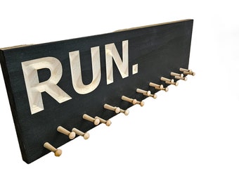 Può includere: Espositore per medaglie in legno nero con la scritta "RUN." in lettere bianche e 18 pioli in legno per appendere le medaglie.