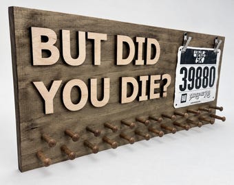 May include: A rectangular wooden display board with a dark brown finish featuring the light-colored wooden text BUT DID YOU DIE? and a metal clip holding a white race bib with the number 39880. The board has multiple wooden pegs.