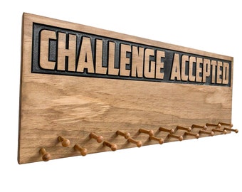 Può includere: Espositore per medaglie da parete in legno con il testo "Challenge Accepted" in lettere nere. L'espositore ha 20 pioli in legno per appendere le medaglie.