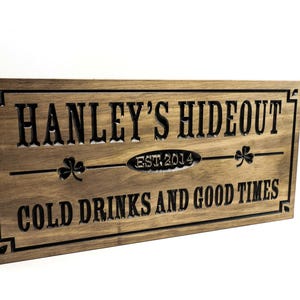 May include: A brown wooden sign with black lettering that reads "Hanley's Hideout Est. 2014 Cold Drinks and Good Times". The sign has a carved shamrock design on each side of the text.