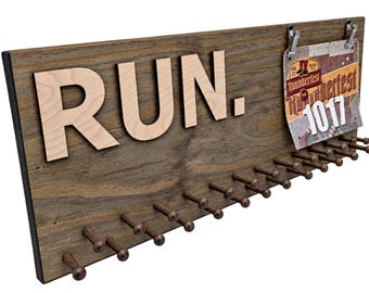 May include: A rectangular wooden medal display board featuring the word RUN. in large light-colored letters. The board includes a row of dark wooden pegs for hanging medals and two metal clips holding a Runtoberfest 5K race bib number 1017.
