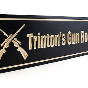 May include: A black wooden sign with a gold border and gold lettering. The sign reads "Trinton's Gun Room" and features two crossed rifles with scopes.