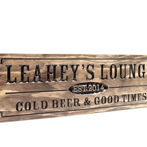 May include: A wooden sign with the text "Leahey's Lounge Est. 2014 Cold Beer & Good Times" in black lettering.