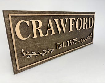 Può includere: Un'insegna in legno marrone scuro con il nome "CRAWFORD" in lettere in rilievo di colore chiaro. L'insegna ha un bordo marrone più chiaro e il testo "Est. 1975". È presente anche un motivo decorativo a foglie.
