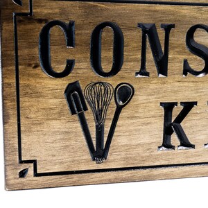 以下が含まれることがあります： 「Constantly in the Kitchen」という文字が刻まれた木製の看板。看板は濃い茶色に着色され、黒い縁取りが施されている。看板には、スパチュラ、泡立て器、スプーンの3つのキッチン用品が刻まれている。