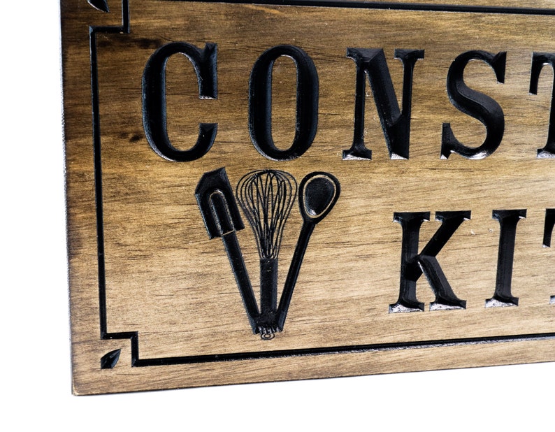 以下が含まれることがあります： 「Constantly in the Kitchen」という文字が刻まれた木製の看板。看板は濃い茶色に着色され、黒い縁取りが施されている。看板には、スパチュラ、泡立て器、スプーンの3つのキッチン用品が刻まれている。