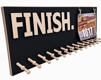May include: A black rectangular wooden display board featuring the word FINISH. in large light-colored wooden letters. The board includes a metal clip holding a race bib labeled Runttoberfest 1017 and a row of light-colored wooden pegs.