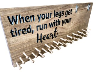Può includere: Tavola espositiva in legno per medaglie con la citazione "When your legs get tired, run with your Heart" intagliata nel legno. La tavola ha piccoli pioli per appendere le medaglie e due clip metalliche in alto.