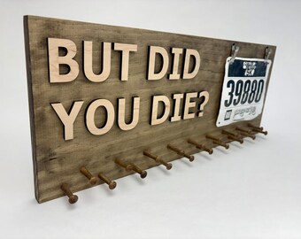 Può includere: Cartello in legno con la scritta "BUT DID YOU DIE?" in lettere di legno chiaro. Il cartello ha una finitura in legno scuro, con piccoli pioli sottostanti per appendere le medaglie. Un pettorale da corsa è agganciato in alto.