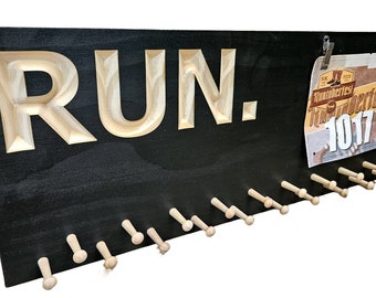 Può includere: Espositore per pettorali da corsa in legno nero con la scritta "RUN." intagliata in legno chiaro. Include una fila di pioli in legno per appendere le medaglie di gara e una clip per esporre un pettorale.