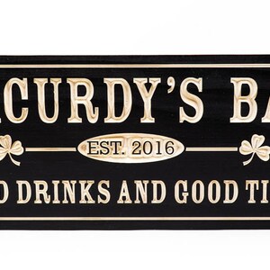 May include: Black wooden sign with gold lettering that reads "Macurdy's Bar Est. 2016 Cold Drinks and Good Times". The sign has two shamrock designs on either side of the text.