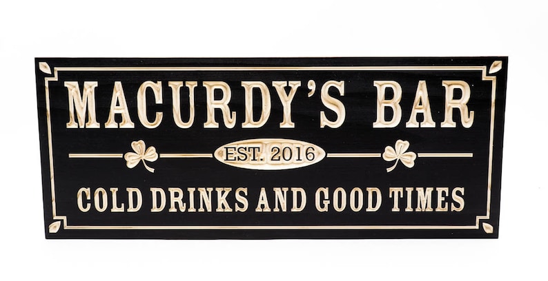 May include: Black wooden sign with gold lettering that reads "Macurdy's Bar Est. 2016 Cold Drinks and Good Times". The sign has two shamrock designs on either side of the text.