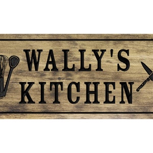以下が含まれることがあります： 黒い文字で「WALLY'S KITCHEN」と書かれた木製の看板。看板は、左側に黒いシルエットのキッチン用品、右側に交差した2本のナイフで飾られています。