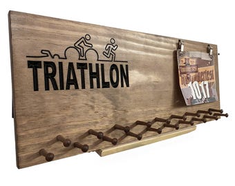 Può includere: Un porta medaglie da parete in legno con ganci per appendere medaglie e pettorali da corsa. Il supporto è a tema triathlon con una silhouette di un triatleta e la parola "TRIATHLON".