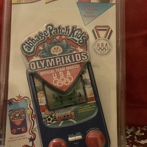 May include: A Cabbage Patch Kids Olympikids handheld soccer game in a blue and red plastic case. The game features a small LCD screen and buttons for kicking, passing, and starting the game. The packaging includes a red, white, and blue medal.