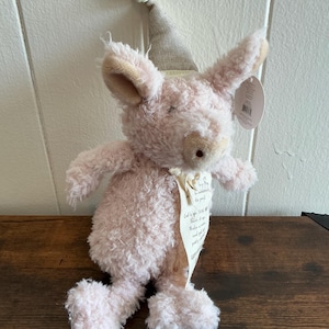 Puede incluir: Un peluche rosa con forma de cerdo, con un gorro gris y un pompón blanco. El cerdo tiene orejas grandes y una etiqueta. Una cinta color crema está atada alrededor de su cuello, con texto impreso. El juguete está sobre una superficie de madera.