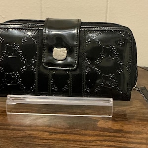May include: Black patent leather wallet with embossed Hello Kitty pattern. The wallet has a snap closure with a silver Hello Kitty emblem and a wrist strap. The wallet is on a clear acrylic stand.