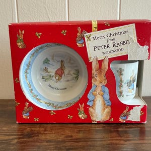 Puede incluir: Un juego de Peter Rabbit Wedgwood en caja. La caja roja presenta ilustraciones de Peter Rabbit y acebo. En el interior, un cuenco y una taza muestran a Peter Rabbit en una escena invernal. La caja dice "Merry Christmas from Peter Rabbit".