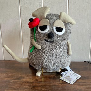 Puede incluir: Un peluche gris con ojos grandes y somnolientos, sosteniendo una flor roja. Tiene orejas color beige y una larga cola beige. El peluche se exhibe sobre una superficie de madera.
