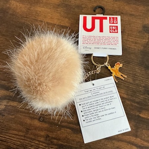 Puede incluir: Un llavero de pompón esponjoso de color beige claro con una cadena dorada y un dije de personaje de Disney. El llavero incluye una etiqueta con el texto "UT" y "Disney Furry Friends".