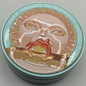 May include: A round, light blue tin featuring a sun-like character with a flame-like creature in its mouth. The tin is labeled "Ghibli Park Valley of Witches" in a circular pattern around the edge. The design is in shades of beige, orange, and brown.
