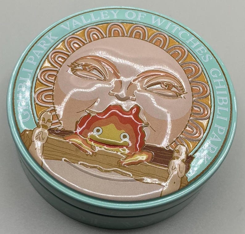 May include: A round, light blue tin featuring a sun-like character with a flame-like creature in its mouth. The tin is labeled "Ghibli Park Valley of Witches" in a circular pattern around the edge. The design is in shades of beige, orange, and brown.