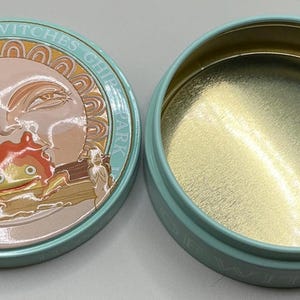 May include: A round, light blue tin with a lid featuring a sun design and the text "GHIBLI PARK VALLEY OF WITCHES". The lid's design includes a cartoon character. The inside of the tin is gold.