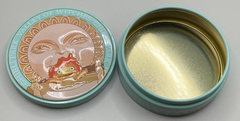 May include: A round, light blue tin with a lid featuring a sun design and the text "GHIBLI PARK VALLEY OF WITCHES". The lid's design includes a cartoon character. The inside of the tin is gold.