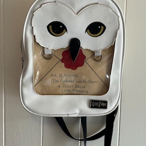 May include: White backpack with an owl design. The top of the bag features an owl face with large, gold-colored eyes and a black beak. A clear pocket displays a letter with the text "MR. H. POTTER, The Cupboard under the Stairs, 4. Privet Drive, Little Whinging."