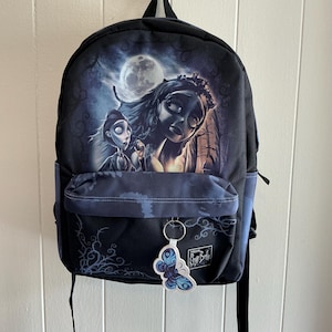 Pode incluir: Mochila preta com uma ilustração gótica de uma noiva e um noivo sob a lua cheia. Possui um bolso frontal e um chaveiro de borboleta. O design é em tons de azul e cinza.