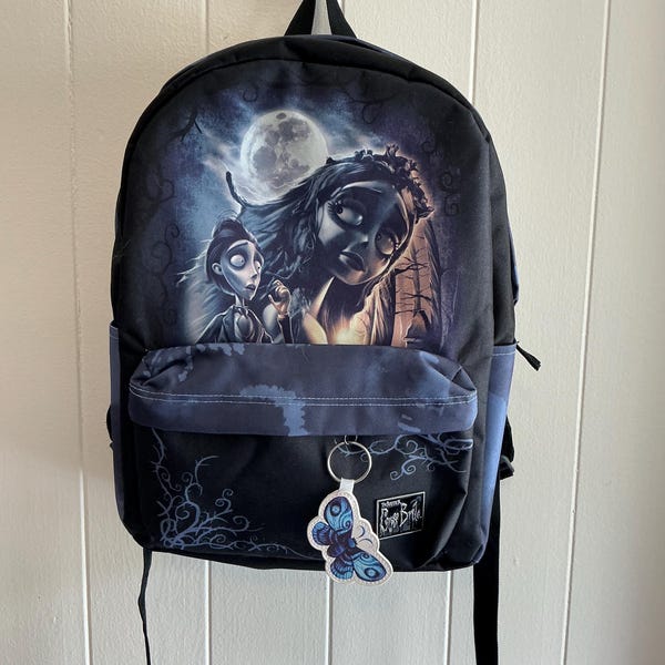 Könnte beinhalten: Schwarzer Rucksack mit einer gotischen Illustration einer Braut und eines Bräutigams unter einem Vollmond. Er hat eine Vordertasche und einen Schmetterlingsschlüsselanhänger. Das Design ist in Blau- und Grautönen gehalten.